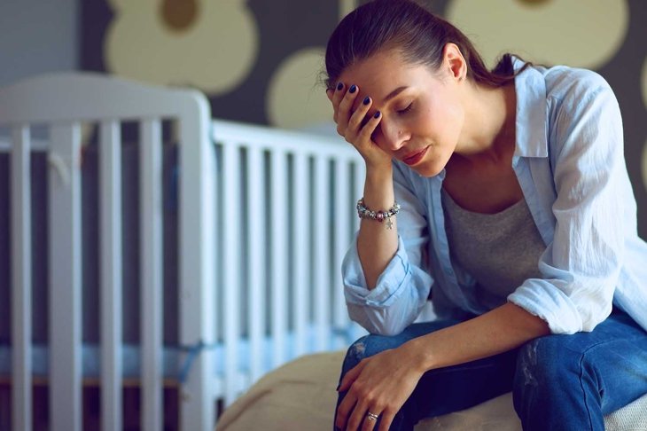 Young tired woman sitting on the bed near children\'s cot.
